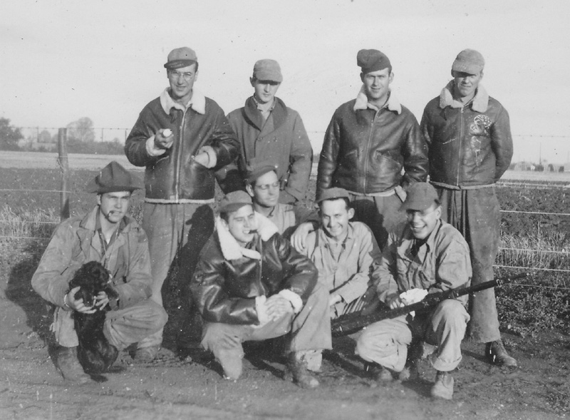 Nine crew men in England with dog mascot and one man holding a rifle.